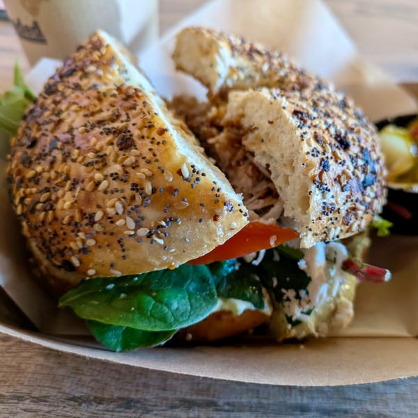 Everything bagel sandwich with chicken, spinach, and tomato, served in a paper tray.