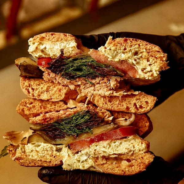 A hand in a black glove holds a large bagel sandwich filled with meat, dill, and vegetables.
