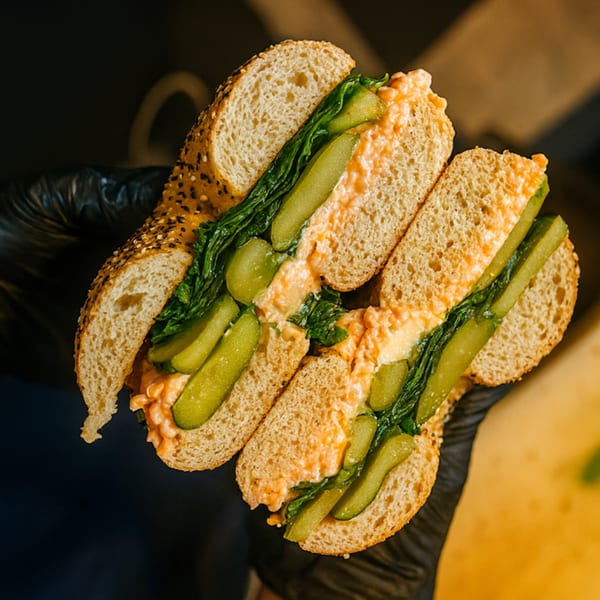 Hands in black gloves holding a bagel sandwich filled with orange spread, green lettuce, and pickles.
