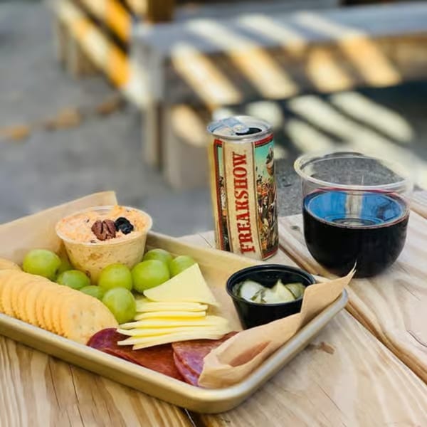 Charcuterie board with cheese, crackers, grapes, dip, and meats, next to a can and a glass of wine.
