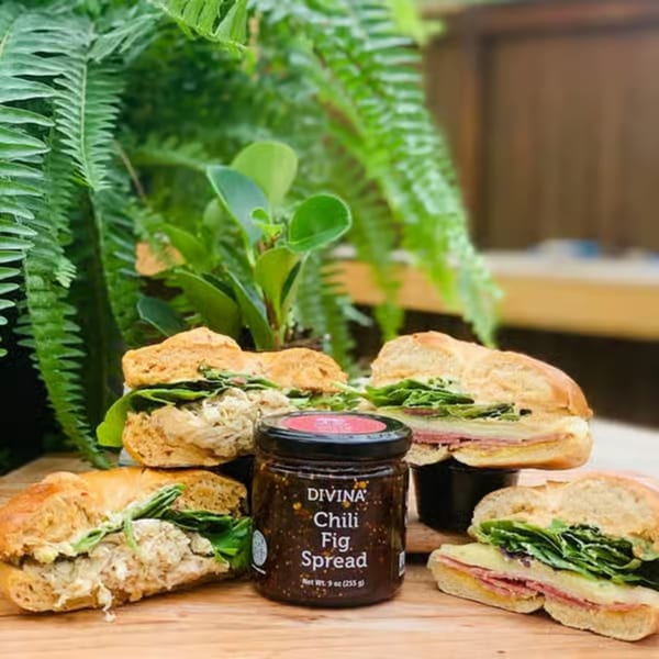 Divina Chili Fig Spread jar surrounded by various sandwiches on a wooden board with green plants.